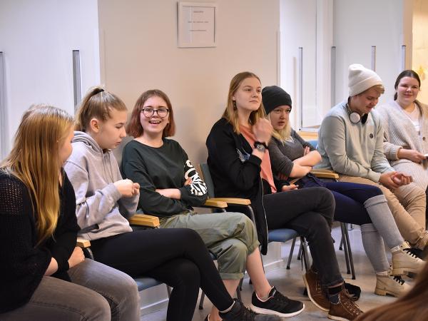 Joukko nuoria istuu tuoleilla seinän vieressä.