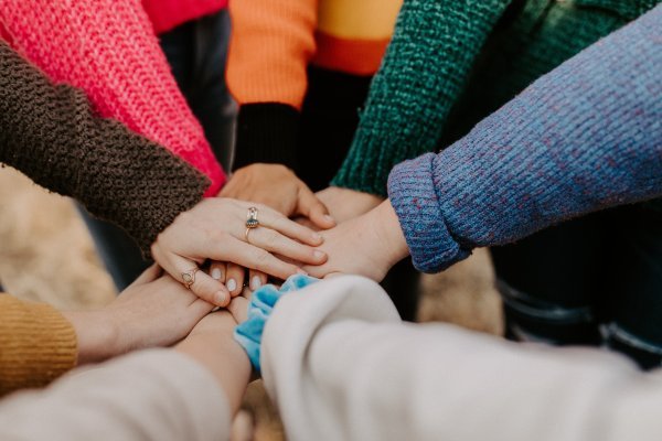 Kuvassa näkyy usean henkilön kädet. Henkilöt seisovat ringissä ja ovat vieneet toisen kätensä keskelle niin, että kädet ovat päällekkäin ja koskettavat toisiaan. Henkilöitä ei muuten näy.